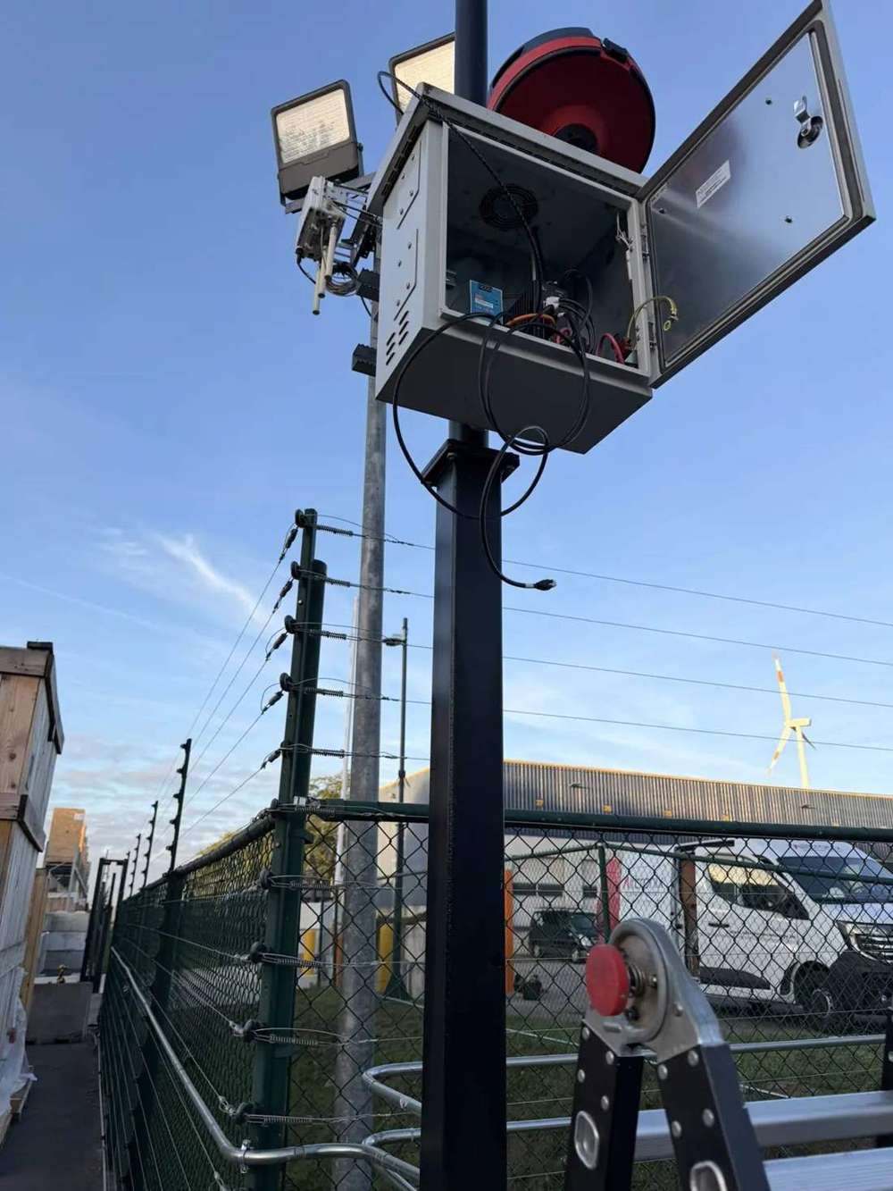 Mast met schijnwerpers, camera en geopende buitenkast met bekabeling naast een omheining op een bedrijfsterrein