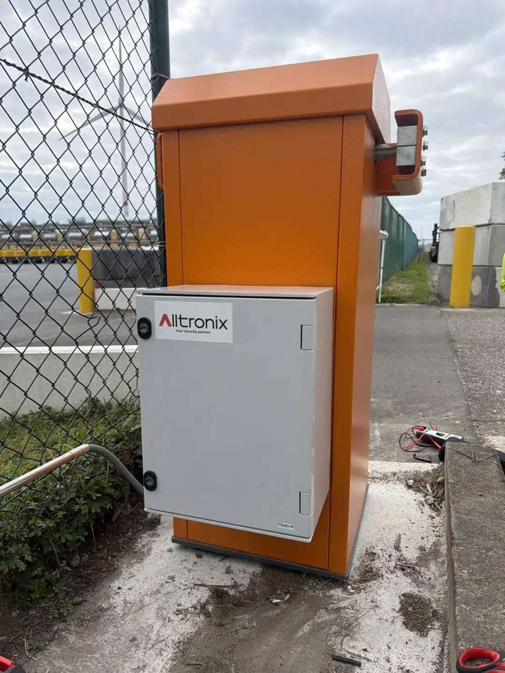 Oranje slagboom met grijze Alltronix-installatiekast aan de toegang van een bedrijfsterrein